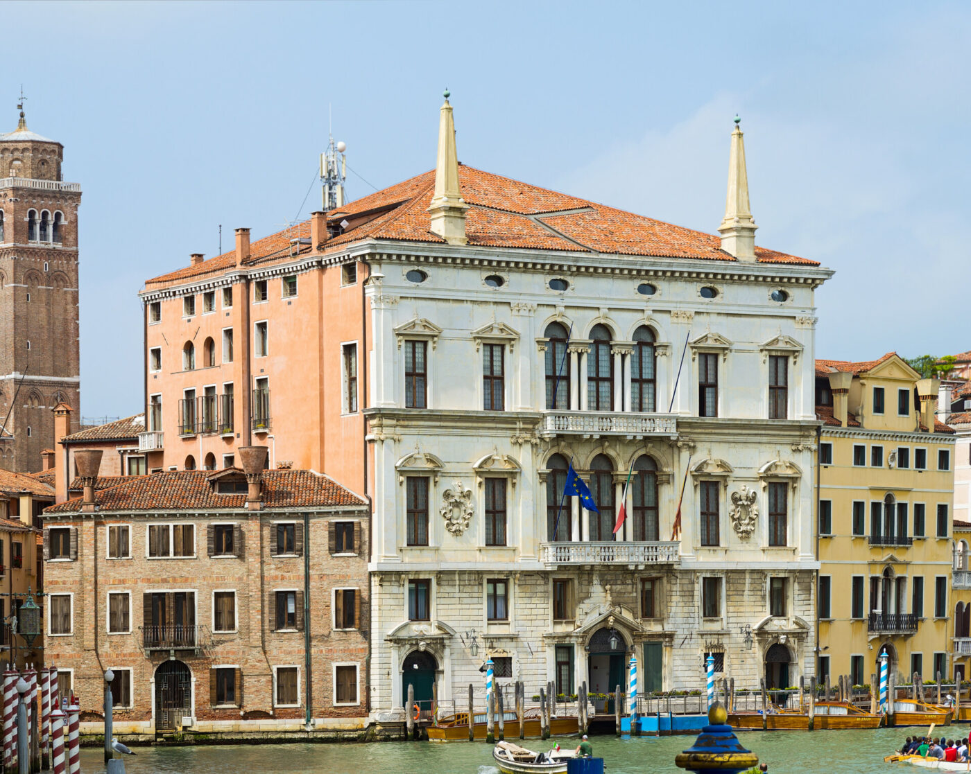 Edificio storico con facciata decorata e tre bandiere su un balcone, affacciato su un canale a Venezia, Italia. Le barche sono ormeggiate lungo l'acqua e sono visibili gli edifici vicini con i tetti in terracotta. Un alto campanile si erge sullo sfondo.