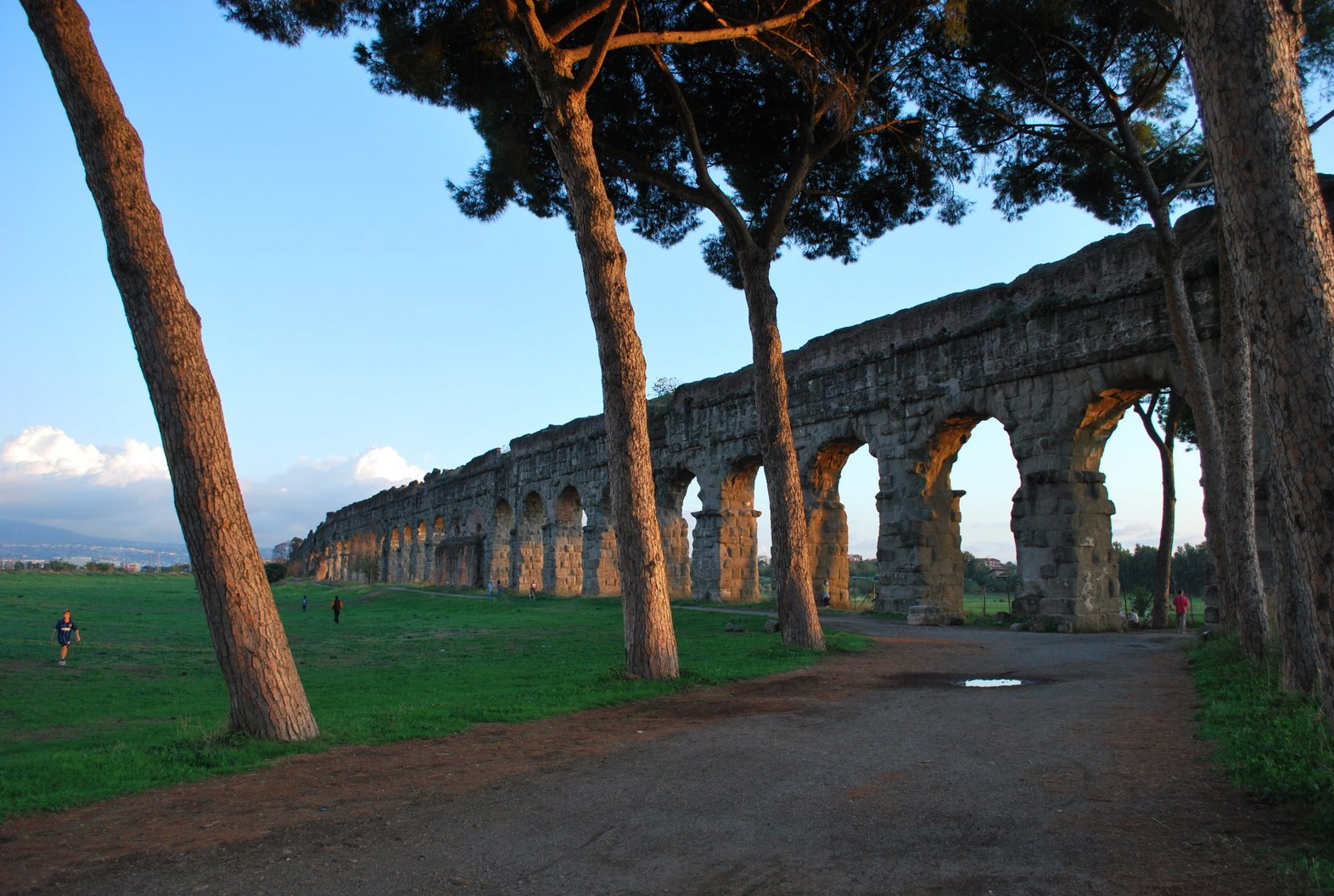 Parco degli Acquedotti - Ancient stone aqueduct arches stretch across a green field, framed by tall pine trees, with blue sky and scattered clouds in the background. A few people walk or stand in the open space.