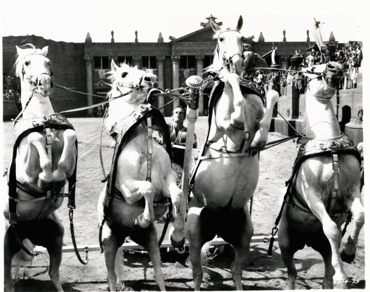 Ben Hur: A dramatic scene of four white horses rearing up while harnessed to a chariot, with a driver holding the reins. Ancient-style buildings and a cheering crowd appear in the background.