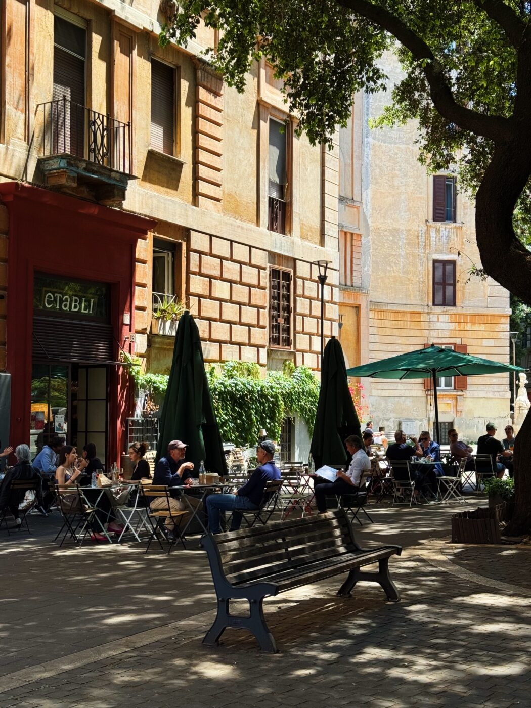 Caffè all'aperto in Piazza Europa - La gente siede ai tavolini all'aperto sotto gli ombrelloni verdi di un caffè in una piazza soleggiata, circondata da vecchi edifici gialli e arancioni. Un grande albero fa ombra e una panchina di legno è visibile in primo piano.