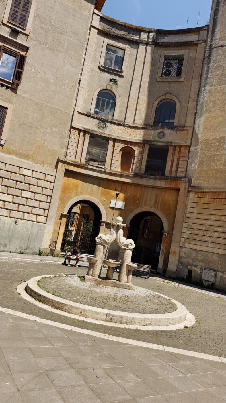 Fontana circolare in pietra in una piccola piazza - Fontana circolare in pietra con sculture astratte in una piccola piazza, circondata da un antico edificio beige curvo con finestre e porte ad arco. Due persone sono sedute su una panchina vicino alla fontana.