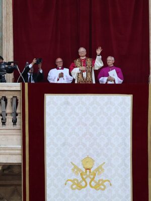 Pope Leo XIV - A religious leader in ornate robes stands on a balcony, waving to a crowd, accompanied by clergy and staff. A large cloth with a gold emblem hangs from the balcony, with cameras and a cross visible nearby.