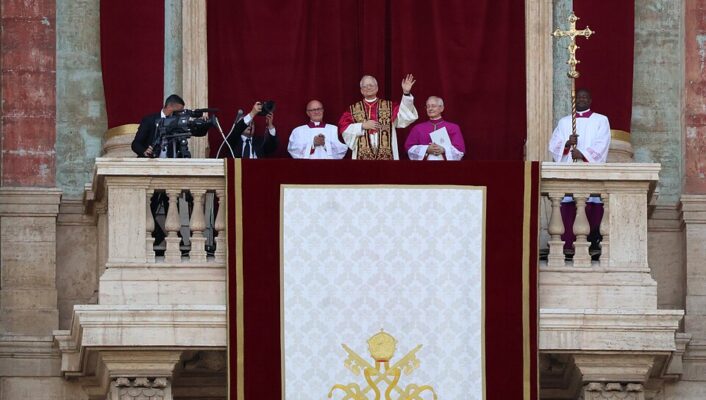Una benedizione papale - Un leader religioso in abiti ornati è in piedi su un balcone, salutando con la mano, affiancato dal clero e dai suoi assistenti. Un grande stendardo con l'emblema papale dorato pende dalla balaustra e due operatori riprendono la scena.