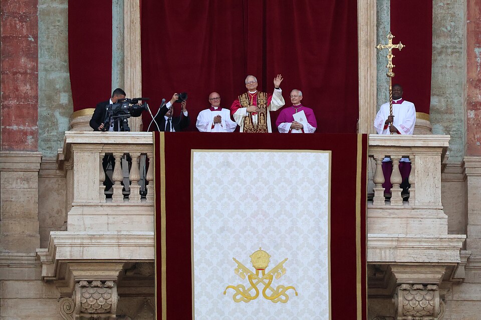 Pope Leo XIV - A religious leader in ornate robes stands on a balcony, waving to a crowd, accompanied by clergy and staff. A large cloth with a gold emblem hangs from the balcony, with cameras and a cross visible nearby.