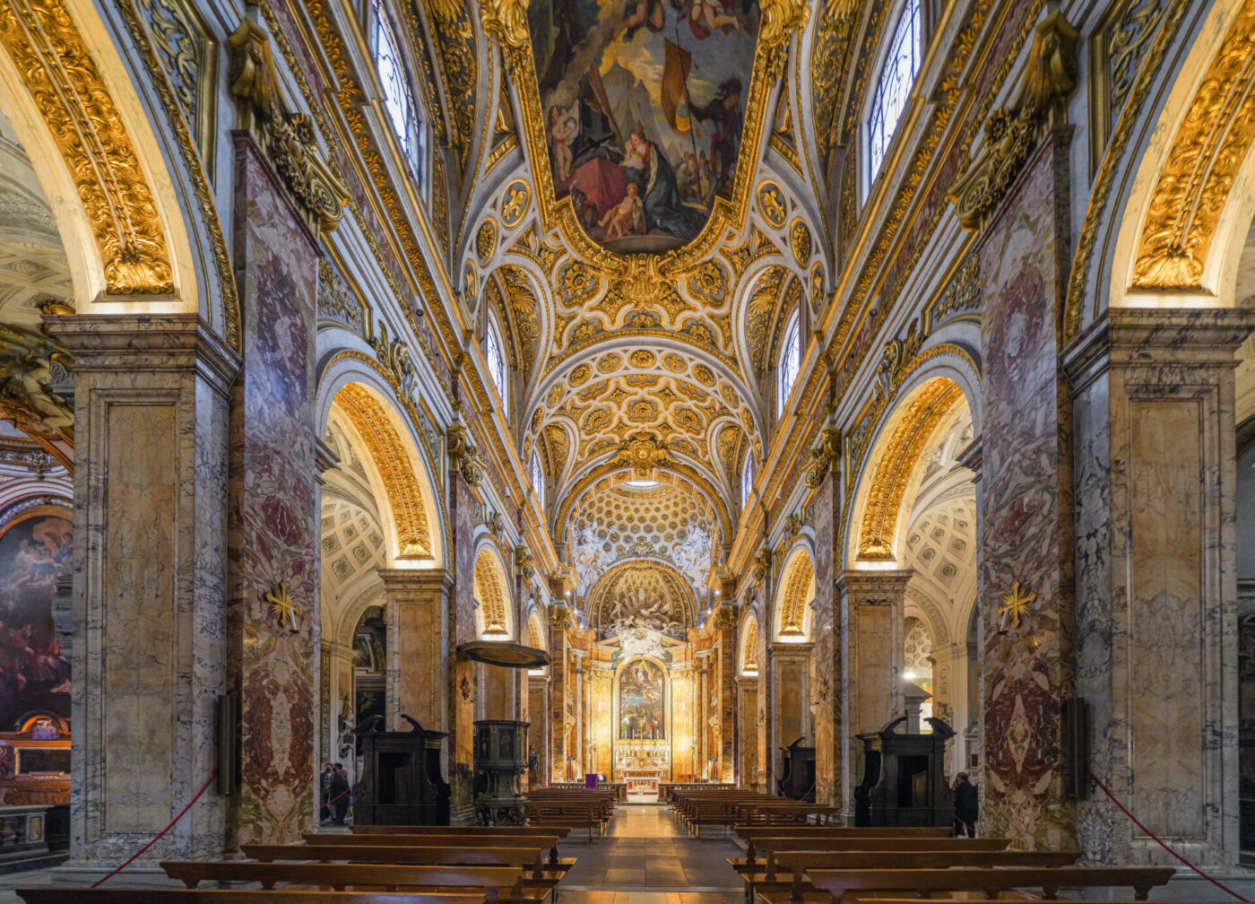 Interno di una grande chiesa con soffitti a volta decorati in oro e bianco, colonne di marmo, panche di legno e un altare decorato in fondo, illuminato da luce naturale e artificiale.