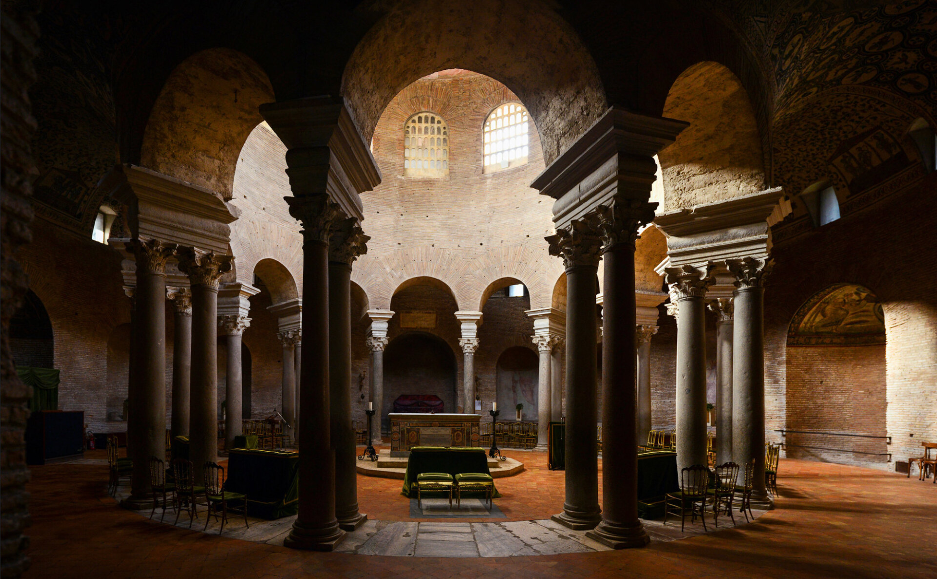 Interior of a round, ancient building with tall stone columns, arched openings, brick walls, and high windows letting in soft light. An altar and benches are arranged in the center on a tiled floor.
