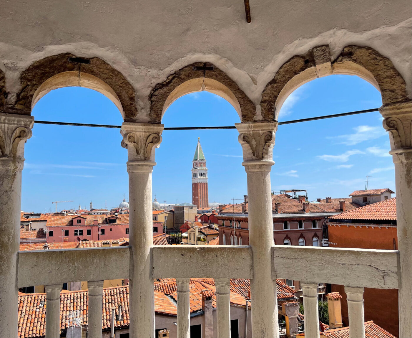 [Nessun titolo dell'immagine fornito] - Vista sui tetti con tegole rosse e su un'alta torre dell'orologio incorniciata da tre archi in pietra, con un cielo azzurro sullo sfondo.