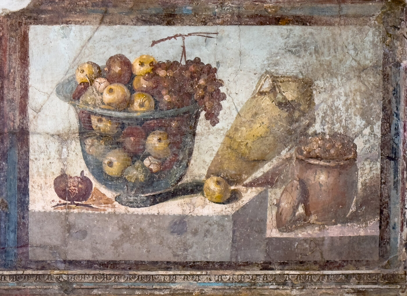 Cesto di mele e uva - Un affresco raffigura un cesto di mele e uva, una brocca a due manici appoggiata su un lato e un piccolo vaso di terracotta pieno di frutta su una superficie di pietra, il tutto su uno sfondo sbiadito e strutturato.