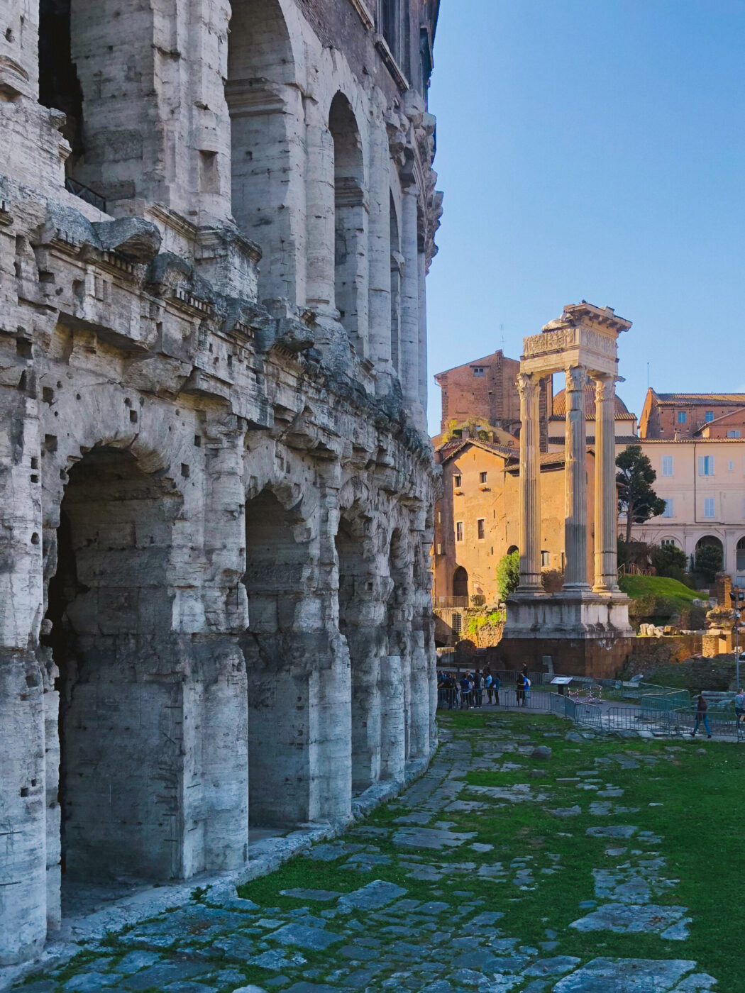 Side view of the ancient Teatro di Marcello's arches in Rome, with sunlit Roman ruins and columns in the background, green grass below, and a group of people walking nearby.