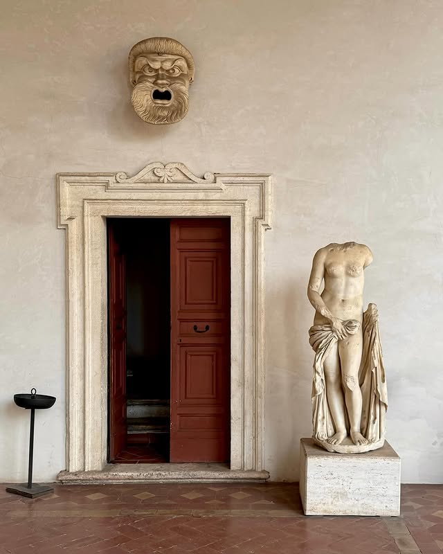 Terme di Diocleziano _ A classical marble statue of a headless male figure stands beside an open red door framed by stone, with a dramatic, expressive stone mask mounted on the wall above the doorway.