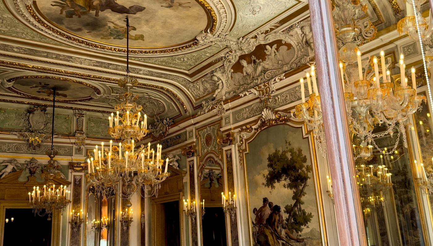 Una sala riccamente decorata con elaborati affreschi sul soffitto, modanature dorate, grandi specchi, lampadari di cristallo e dipinti classici alle pareti, illuminata da calde luci simili a quelle delle candele.