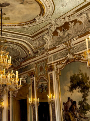 Una sala riccamente decorata con elaborati affreschi sul soffitto, modanature dorate, grandi specchi, lampadari di cristallo e dipinti classici alle pareti, illuminata da calde luci simili a quelle delle candele.