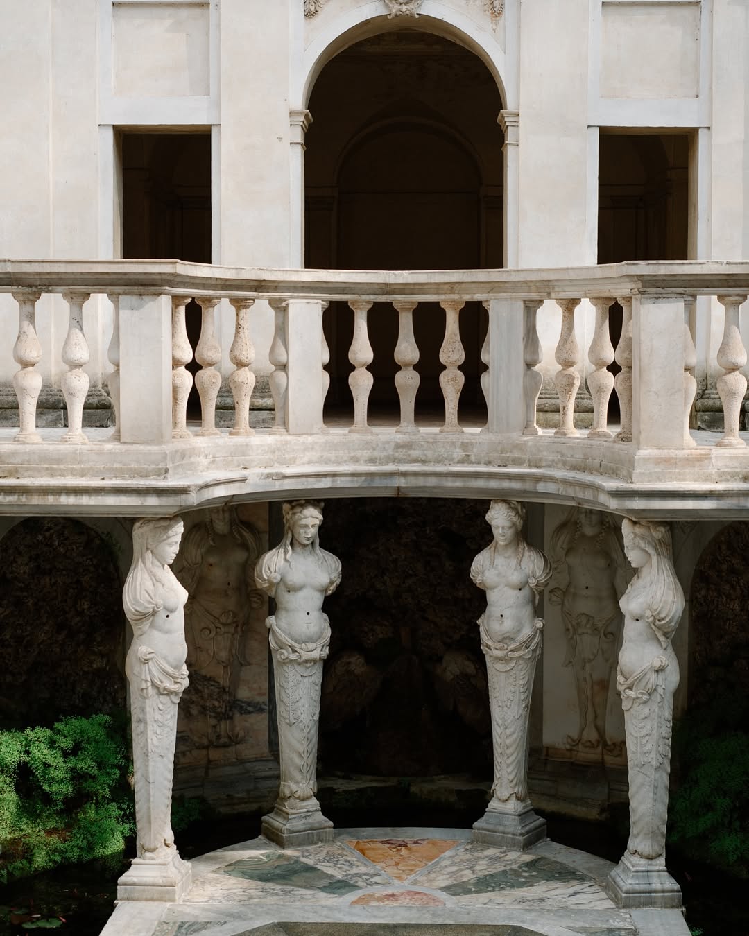 Villa Giulia _ A two-level stone structure with caryatid figures - A two-level stone structure with a balustrade balcony overlooks three sculpted caryatid figures supporting the upper level. Lush green plants surround the lower area, and an arched entrance is visible above.