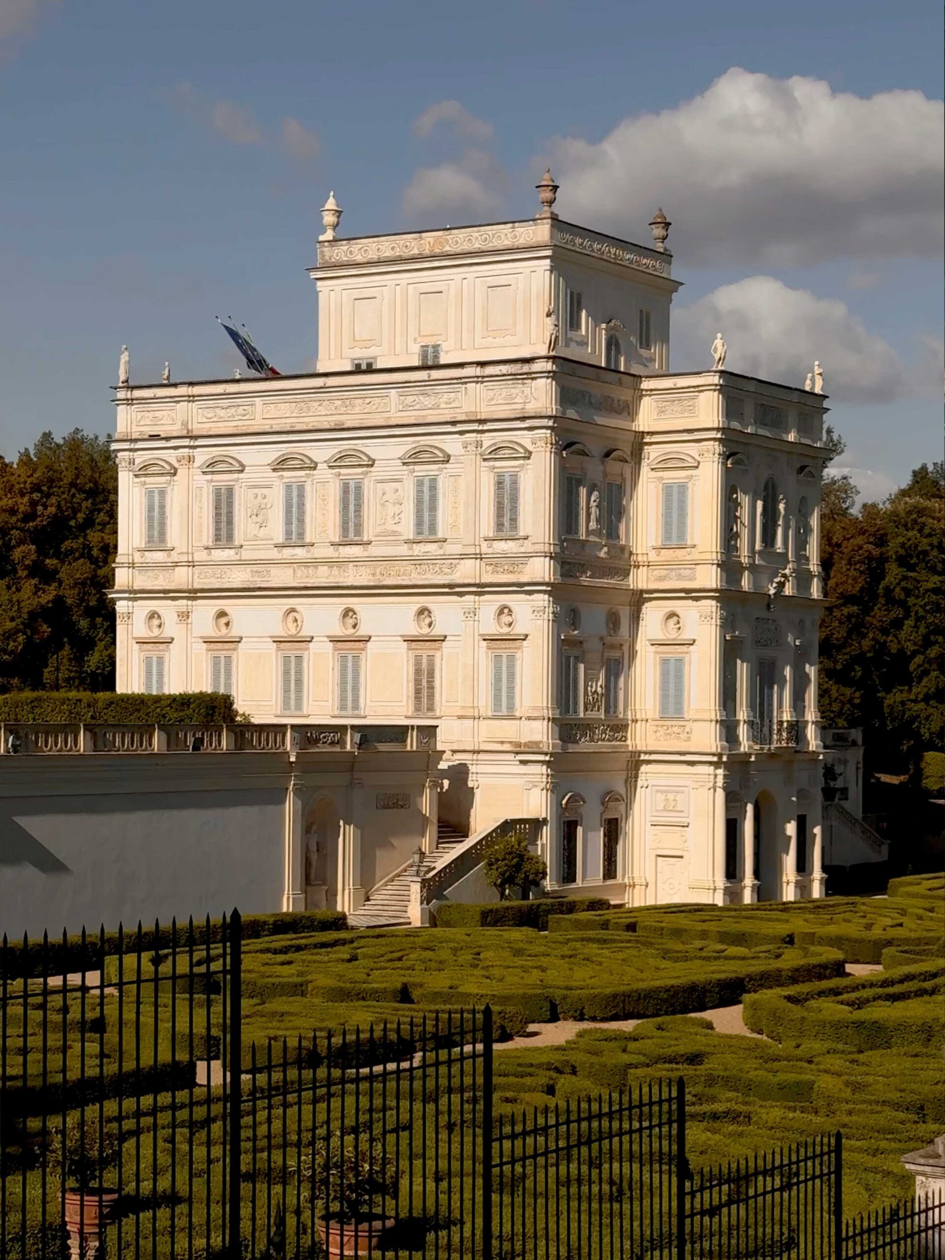 Villa Doria Pamphilji - A grand, light-colored neoclassical building stands among manicured hedges and gardens, with a partly cloudy sky and trees in the background. Ornate details decorate the façade and wrought iron fencing lines the foreground.