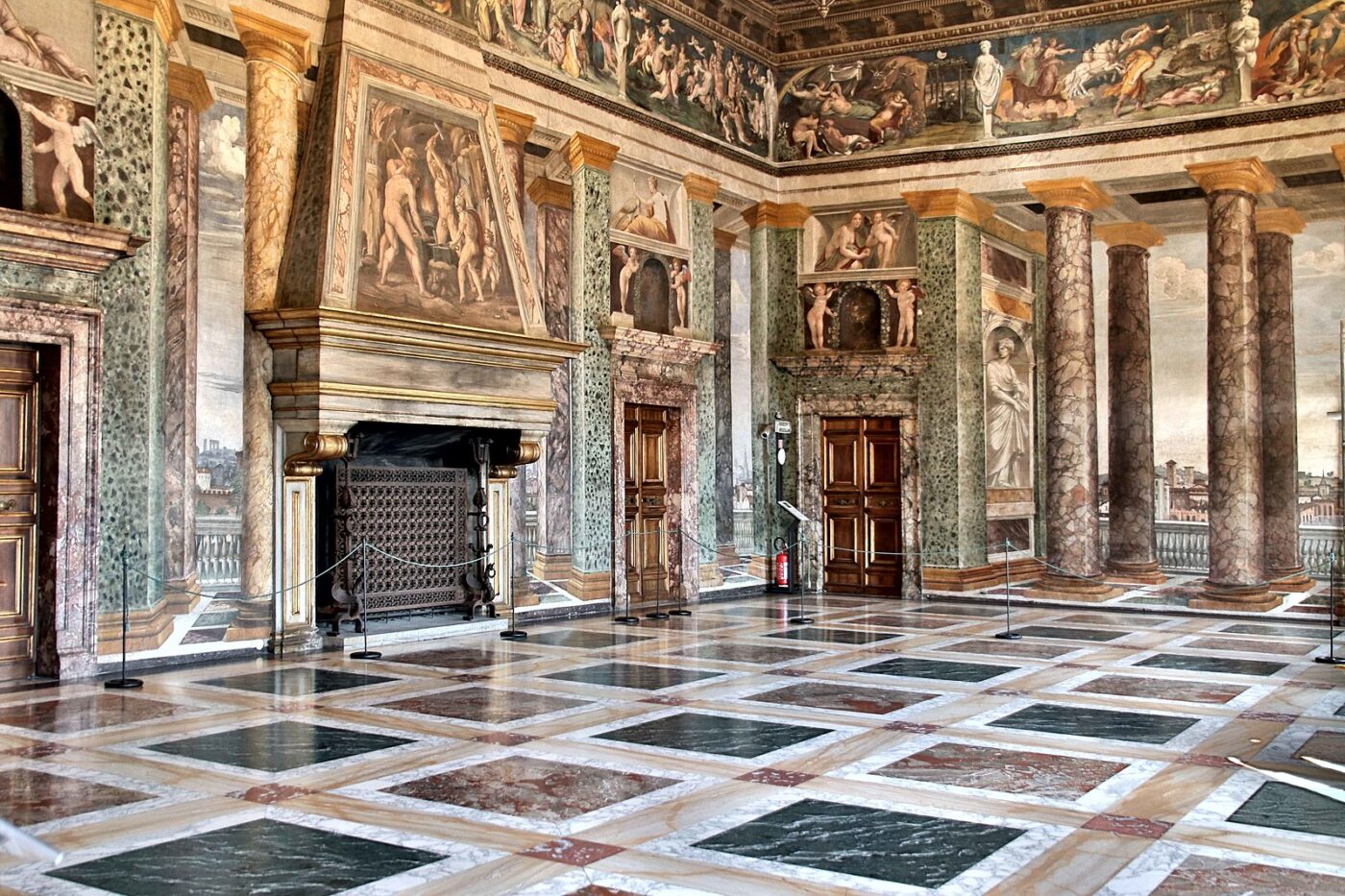 Villa Farnesina_A grand, ornate hall with marble floors, tall marble columns, detailed wall and ceiling frescoes, and large wooden doors, illuminated by natural light. The room is empty and richly decorated in a classical style.