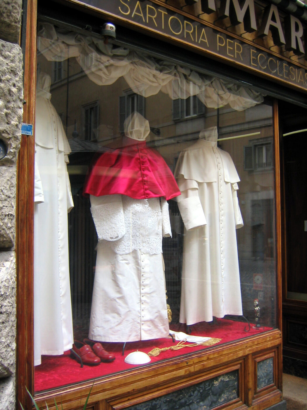 Sartoria per Ecclesiastici - Una vetrina espone abiti ecclesiastici su misura, tra cui vesti bianche, un mantello rosso con le spalle, un cappello bianco e scarpe rosse; un'insegna in italiano sopra recita "Sartoria per Ecclesiastici.