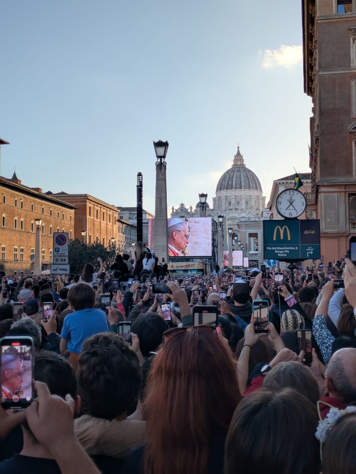 St. Peter’s Square Crowd - A large crowd gathers in St. Peter’s Square, Vatican City, many holding up phones to capture a screen showing the pope. St. Peter’s Basilica and a McDonald’s sign are visible in the background.