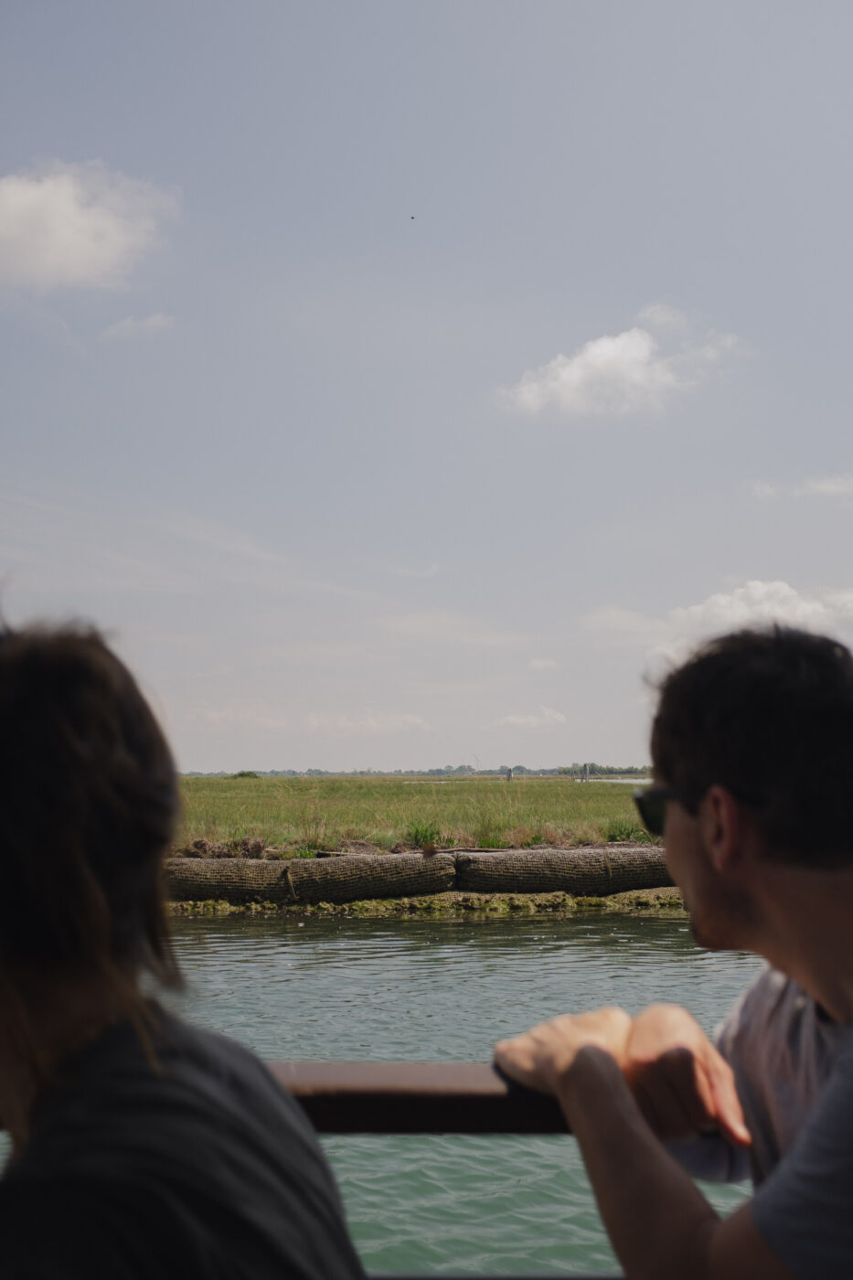 [Nessun titolo dell'immagine] - Due persone su una barca osservano un paesaggio erboso e l'acqua sotto un cielo parzialmente nuvoloso; una persona è in primo piano, l'altra è più indietro, entrambe guardano verso la riva.