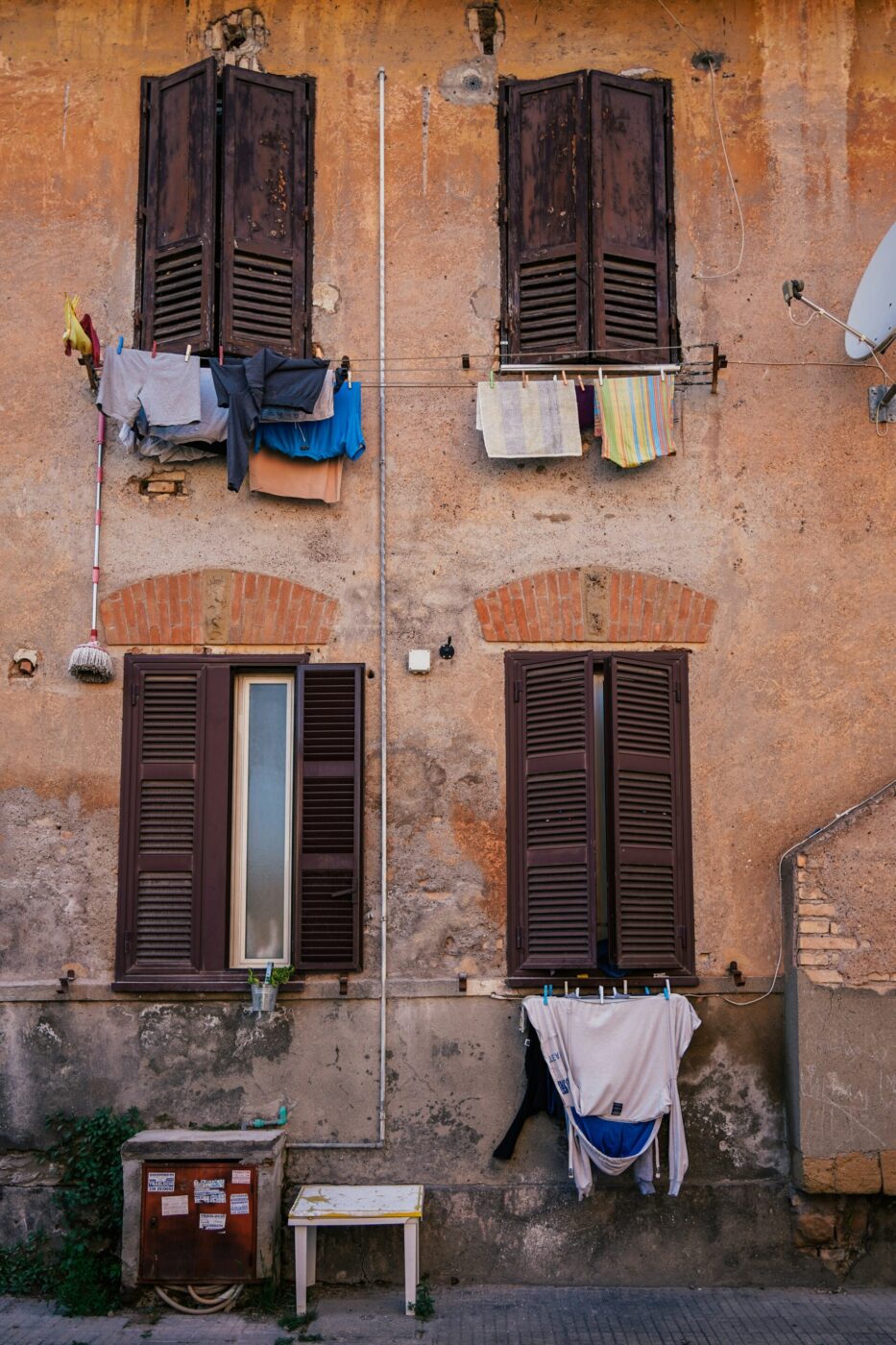 An old, weathered building with four brown shuttered windows; laundry hangs on clotheslines beneath each window. The wall is stained and rustic, with a satellite dish and outdoor pipes visible.