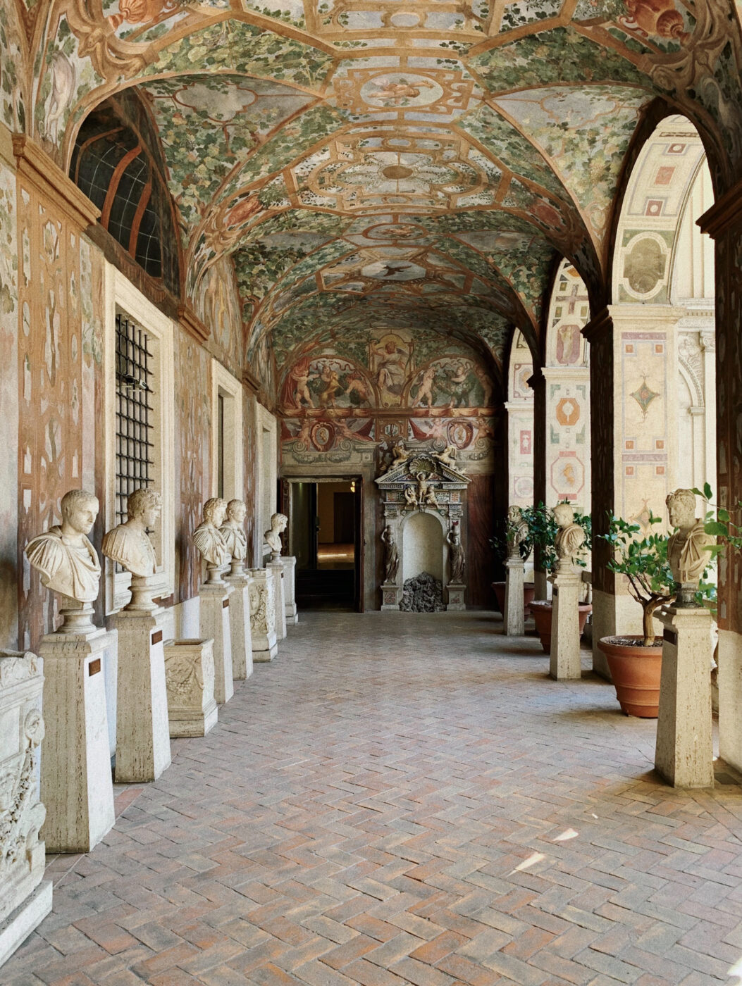 Palazzo Altemps - A corridor with a vaulted, ornately painted ceiling lined with classical marble busts on pedestals; the walls and ceiling are decorated with frescoes, and potted plants are placed along the right side.