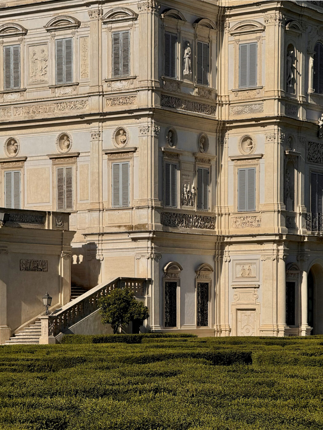 Villa Doria Pamphilj with detailed stonework, sculptures, and closed gray shutters stands behind manicured green hedges and a staircase with a stone railing. Sunlight highlights the building’s classic architecture.