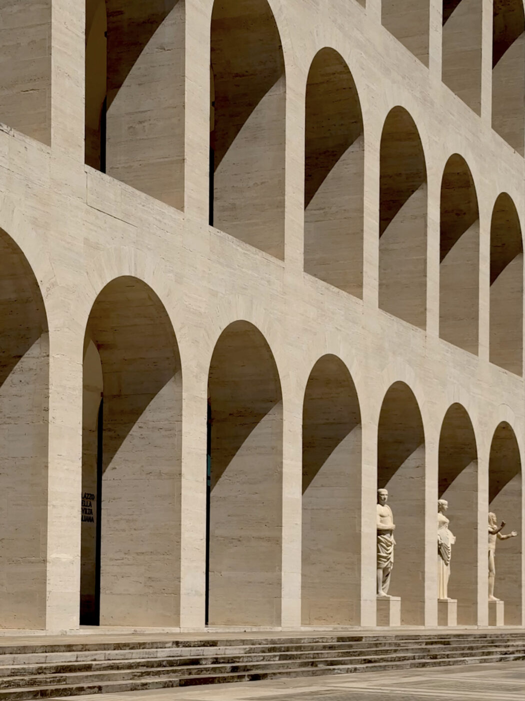 Palazzo della Civiltà Italiana: A stone building with multiple rows of large archways. Three classical statues stand between the arches on a lower level, casting long shadows in bright sunlight.