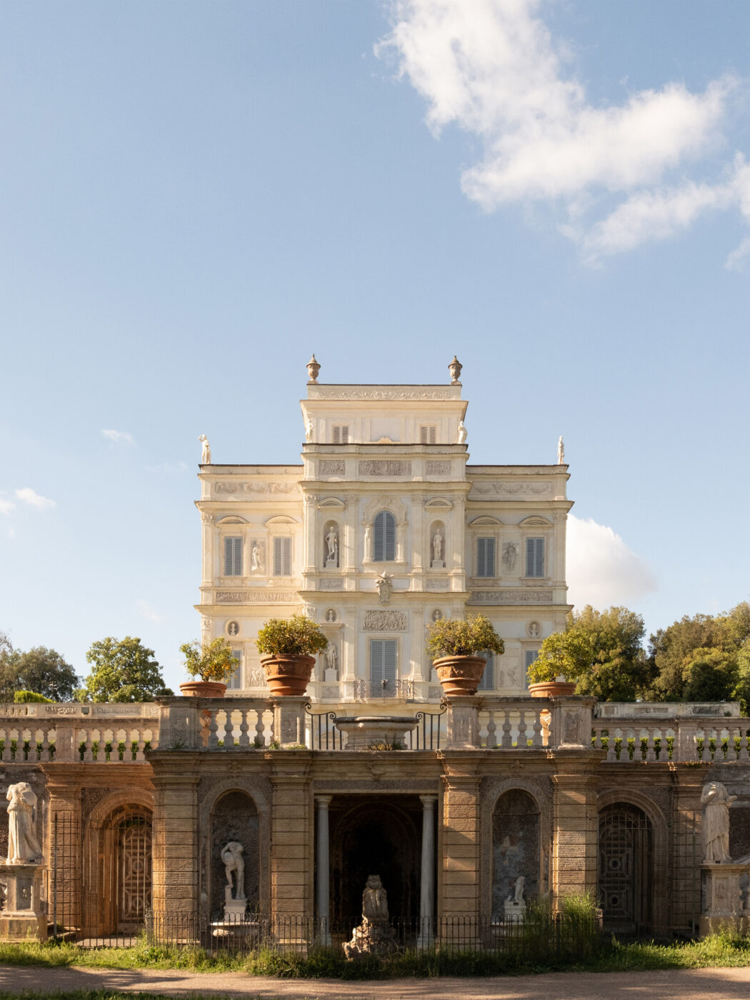 Villa Doria Pamphilj - A grand classical villa with ornate white façade stands behind a stone terrace adorned with statues, columns, and large potted plants, set against a blue sky with scattered clouds.