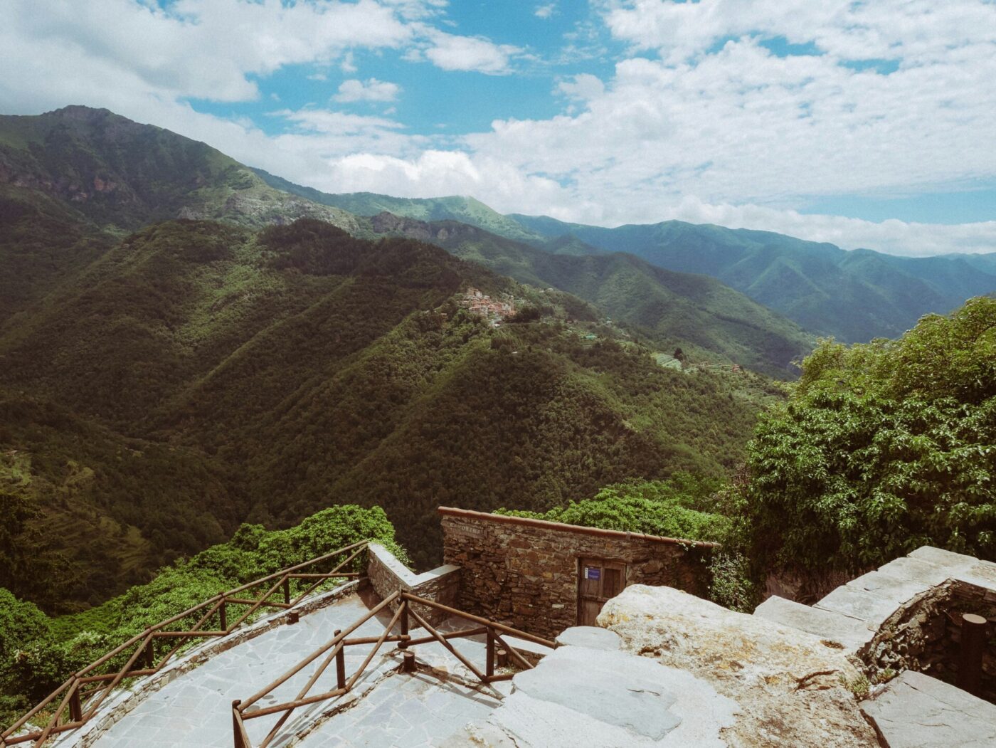 Paesaggio montano panoramico - Un paesaggio montano panoramico con lussureggianti colline verdi sotto un cielo parzialmente nuvoloso, osservato da una terrazza in pietra con una ringhiera in legno e gradini in pietra in primo piano.