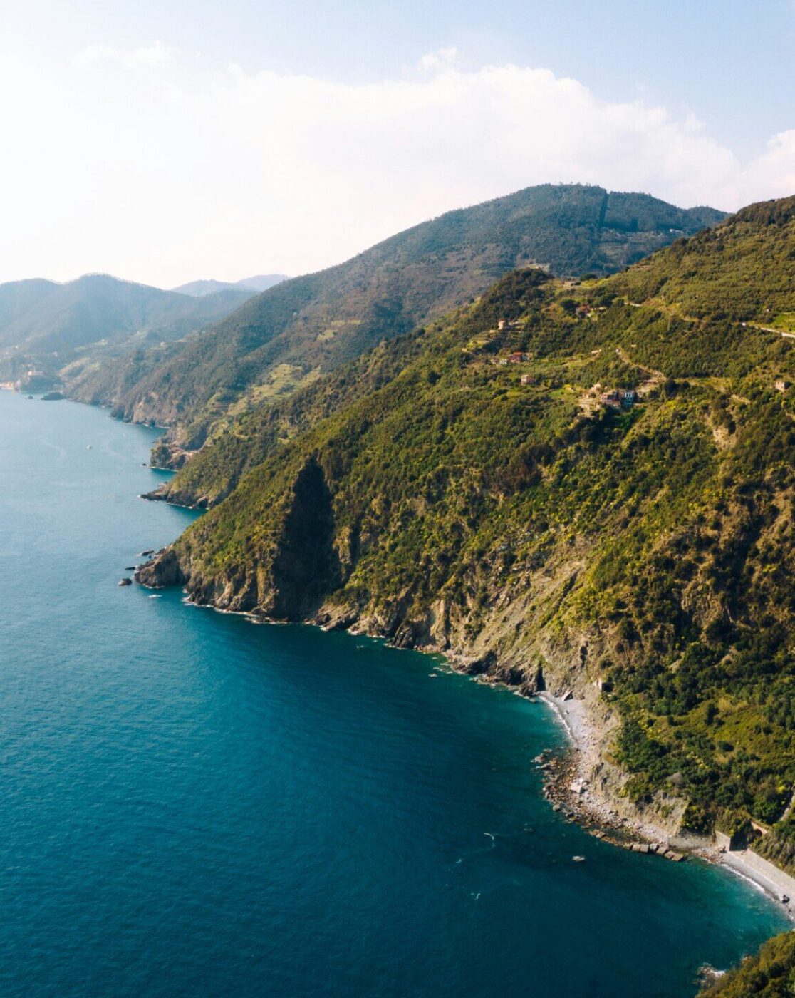 Aerial view of a rugged coastline with green hills meeting clear blue sea, small beaches nestled between cliffs, and distant mountains under a bright, sunny sky.