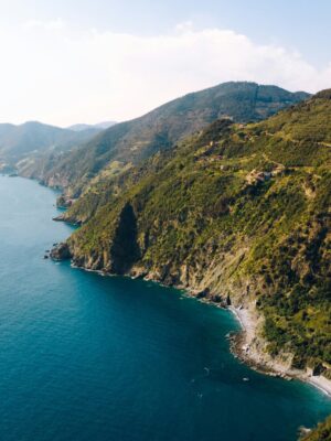 Aerial view of a rugged coastline with green hills meeting clear blue sea, small beaches nestled between cliffs, and distant mountains under a bright, sunny sky.