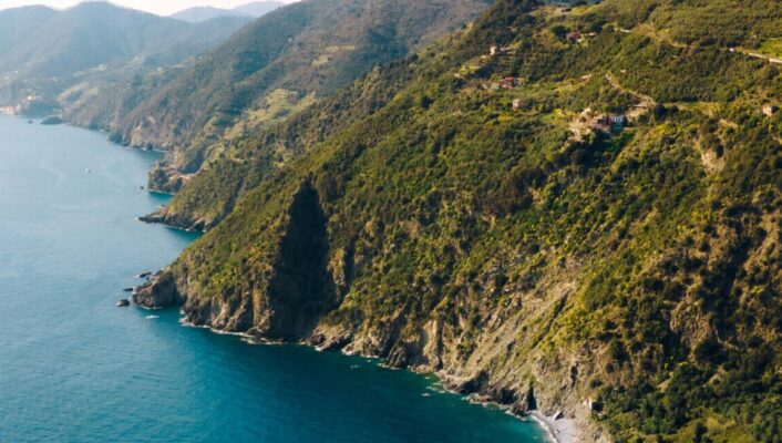 Aerial view of a rugged coastline with green hills meeting clear blue sea, small beaches nestled between cliffs, and distant mountains under a bright, sunny sky.