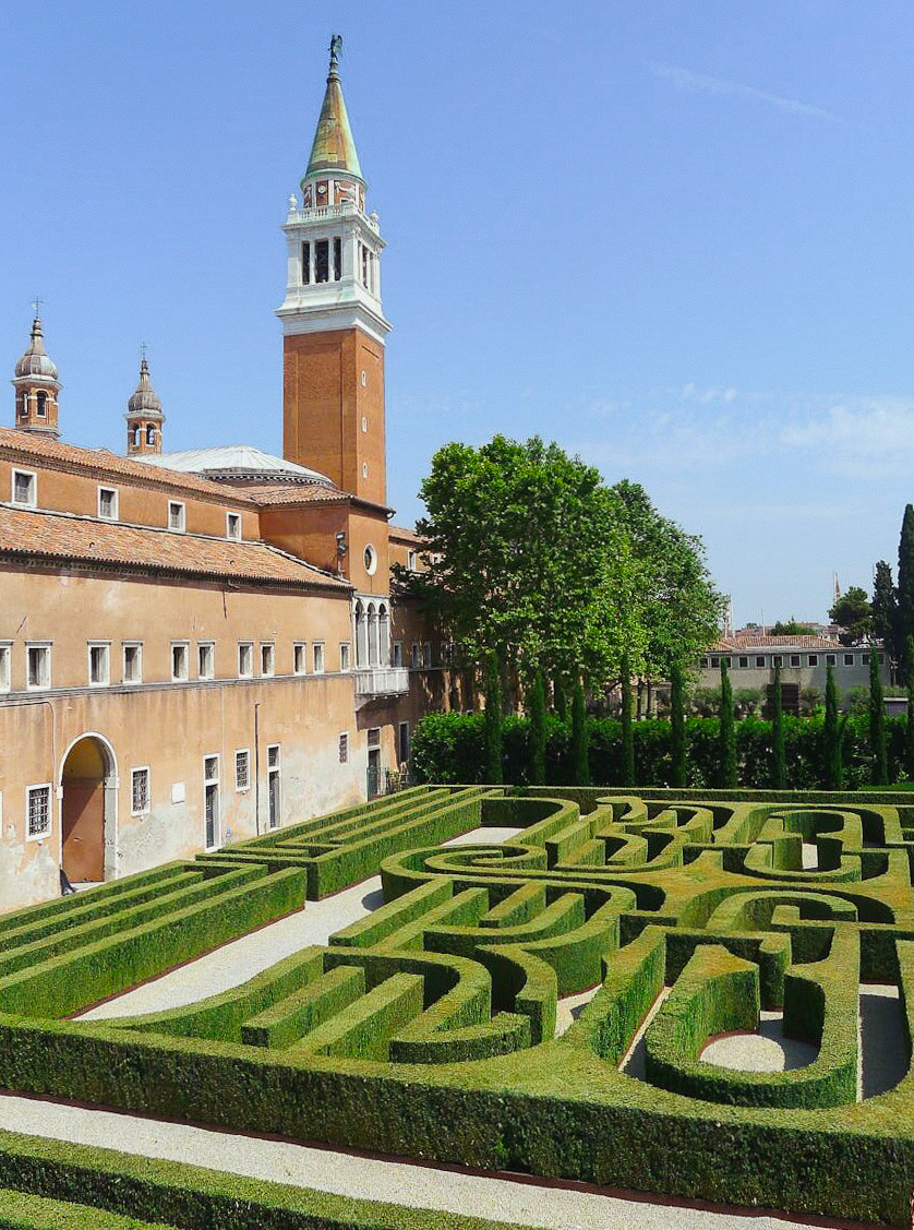 Un labirinto di siepi ben curato si trova accanto a un edificio storico con finestre ad arco e un alto campanile, sotto un cielo azzurro e limpido. Alberi rigogliosi costeggiano il giardino, creando un'atmosfera di pace.