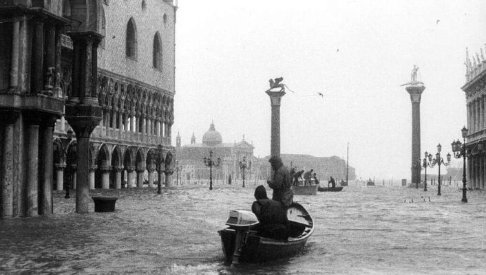 St. Mark’s Square Flooding - A black-and-white photo of people in a small boat navigating a heavily flooded St. Mark’s Square in Venice, Italy. Historic buildings and columns are partially submerged under high water.