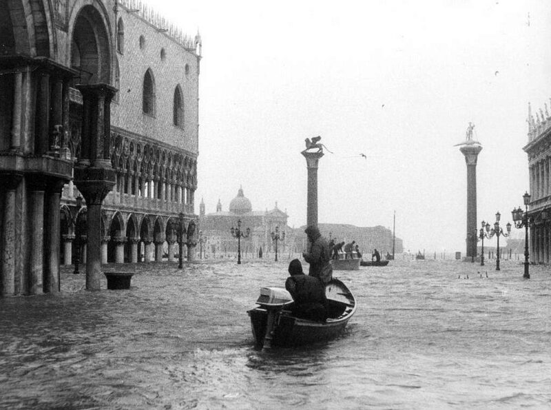 St. Mark’s Square Flooding - A black-and-white photo of people in a small boat navigating a heavily flooded St. Mark’s Square in Venice, Italy. Historic buildings and columns are partially submerged under high water.