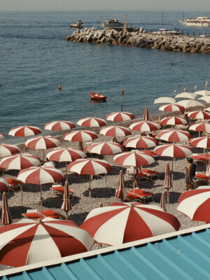 Una scena di spiaggia con file di ombrelloni rossi e bianchi su ciottoli, affacciati sul mare calmo - Testo Alt: Una scena di spiaggia con file di ombrelloni rossi e bianchi disposti ordinatamente su una spiaggia di ciottoli. Gli ombrelloni si affacciano sul mare tranquillo e blu. Sullo sfondo, alcune barche sono attraccate a un molo roccioso. In primo piano è visibile parte di un tetto turchese.