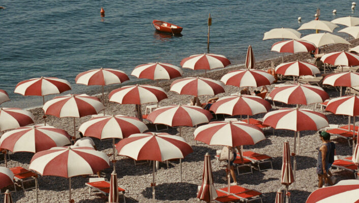 Rows of red and white beach umbrellas cover a pebbled shore by the sea, with a turquoise roof in the foreground and boats and a stone pier in the background.