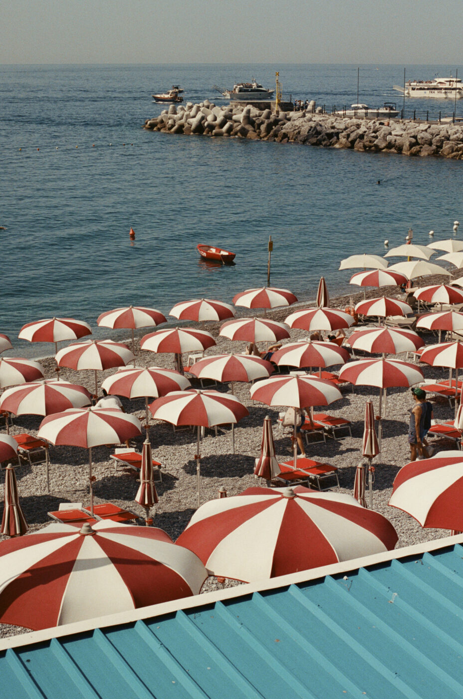 Una scena di spiaggia con file di ombrelloni rossi e bianchi su ciottoli, affacciati sul mare calmo - Testo Alt: Una scena di spiaggia con file di ombrelloni rossi e bianchi disposti ordinatamente su una spiaggia di ciottoli. Gli ombrelloni si affacciano sul mare tranquillo e blu. Sullo sfondo, alcune barche sono attraccate a un molo roccioso. In primo piano è visibile parte di un tetto turchese.