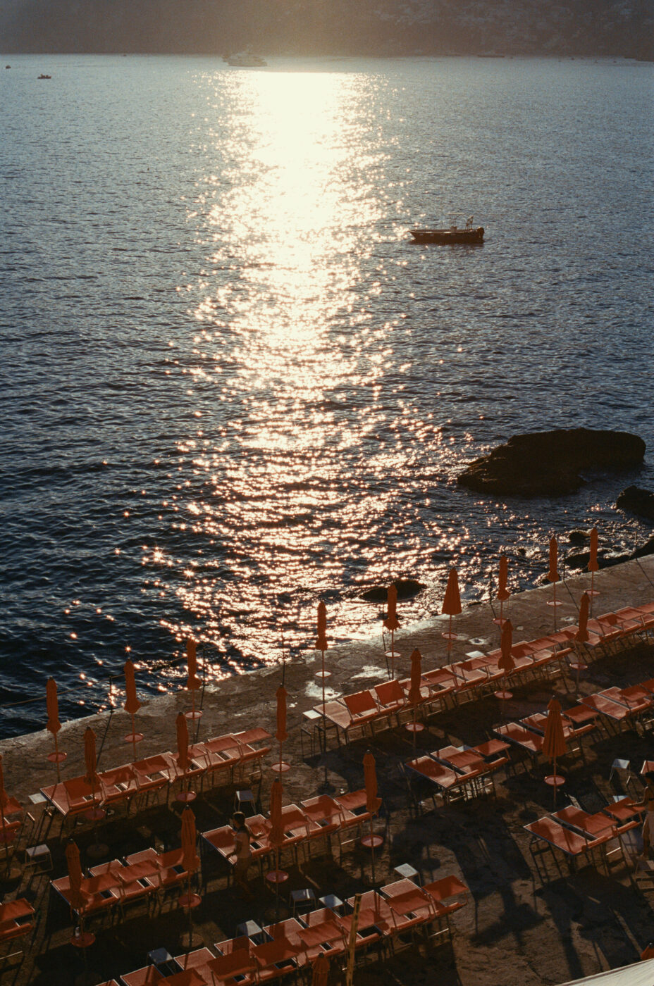 File di sedie a sdraio e ombrelloni arancioni vuoti - File di sedie a sdraio e ombrelloni arancioni vuoti costeggiano il lungomare mentre il sole si riflette sul mare calmo, con una piccola barca che galleggia in lontananza e una costa rocciosa visibile.