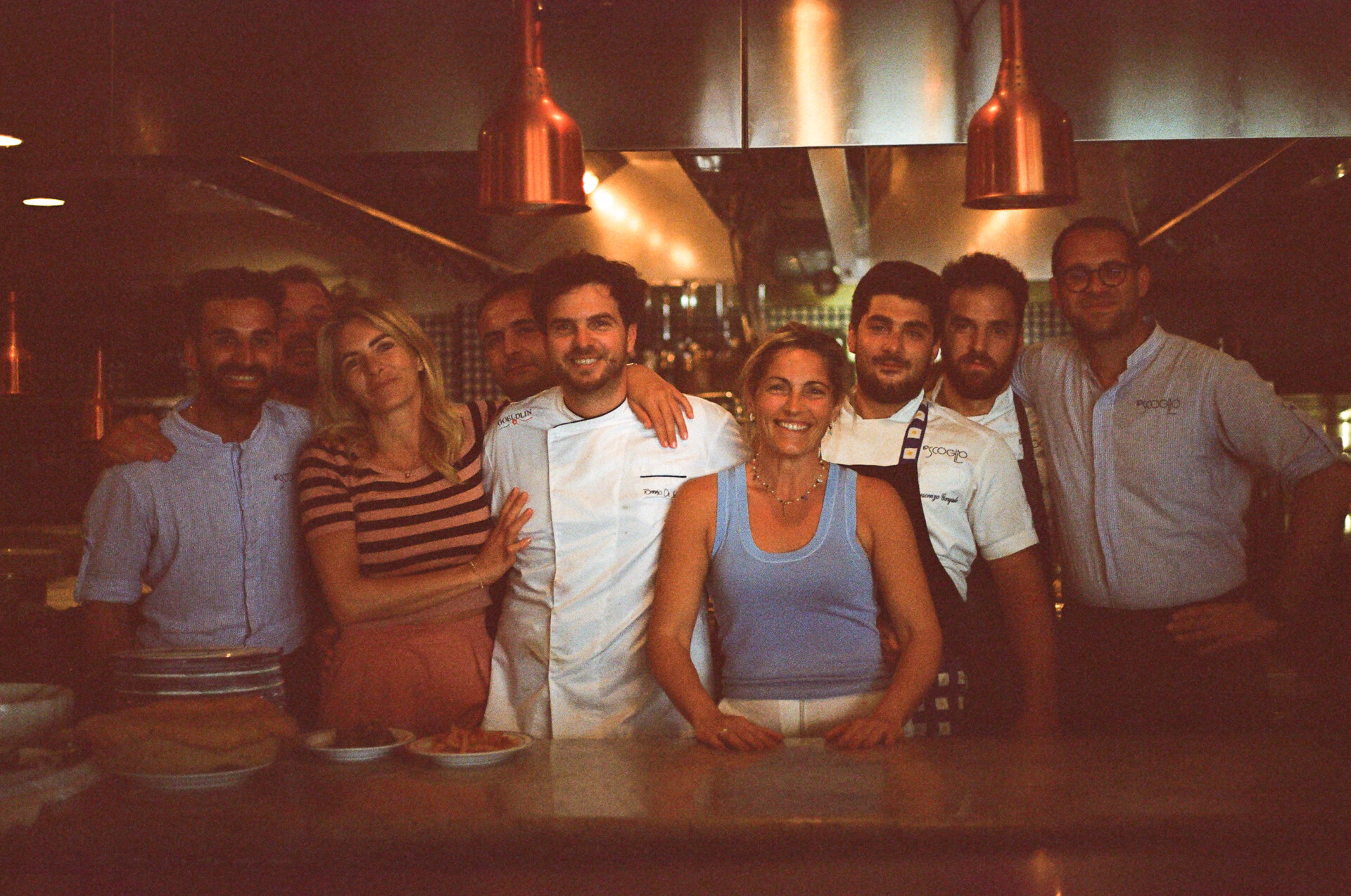 Il testo alt deve essere formattato come segue: [Un gruppo di otto persone, tra cui cuochi in uniforme bianca e altri in abiti casual, posano e sorridono insieme dietro il bancone di un ristorante in una cucina caldamente illuminata. Quindi, se il titolo dell'immagine è "Team Celebration", il testo alt completo sarà: Team Celebration - Un gruppo di otto persone, tra cui cuochi in uniforme bianca e altri in abiti casual, posano e sorridono insieme dietro il bancone di un ristorante in una cucina ben illuminata.