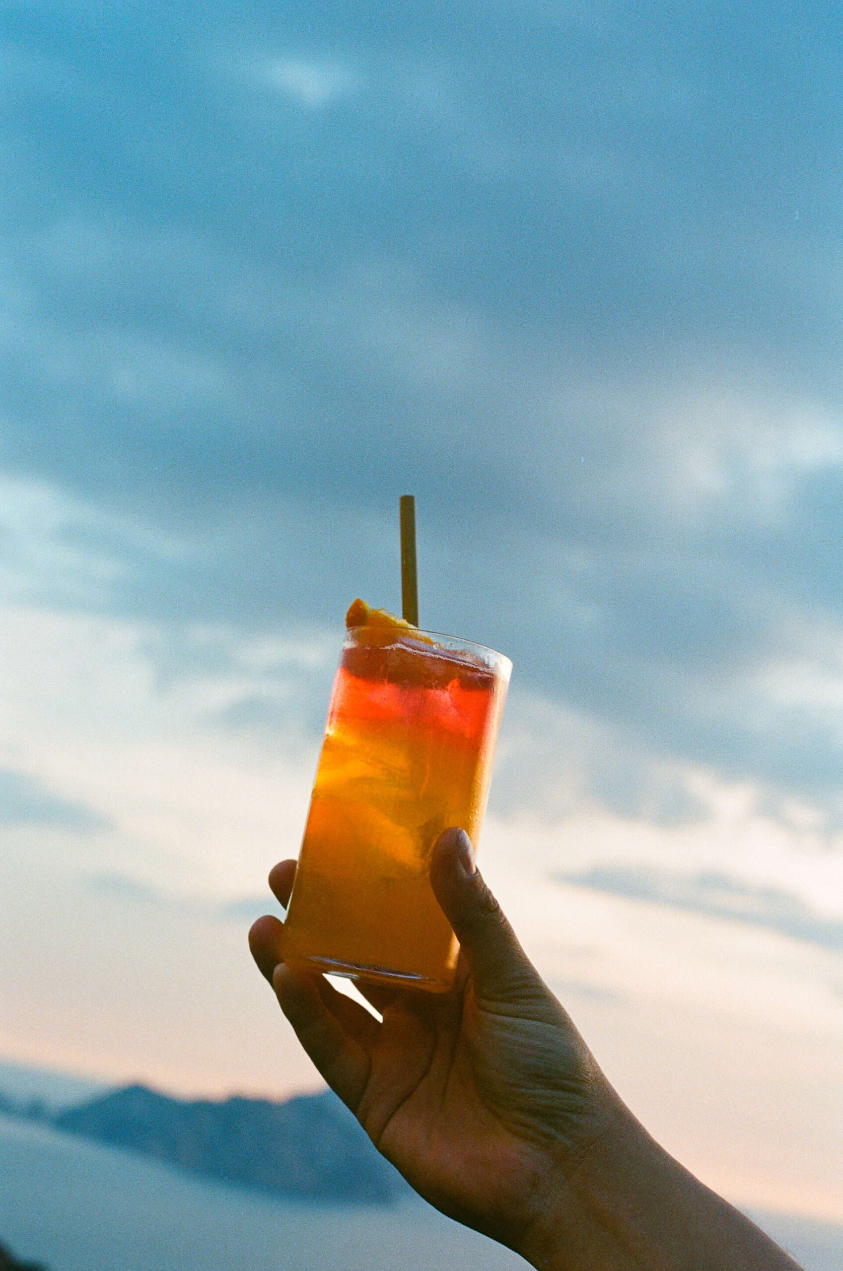Una mano regge un cocktail colorato guarnito con una fetta d'arancia e una cannuccia in un cielo nuvoloso con montagne lontane sullo sfondo.