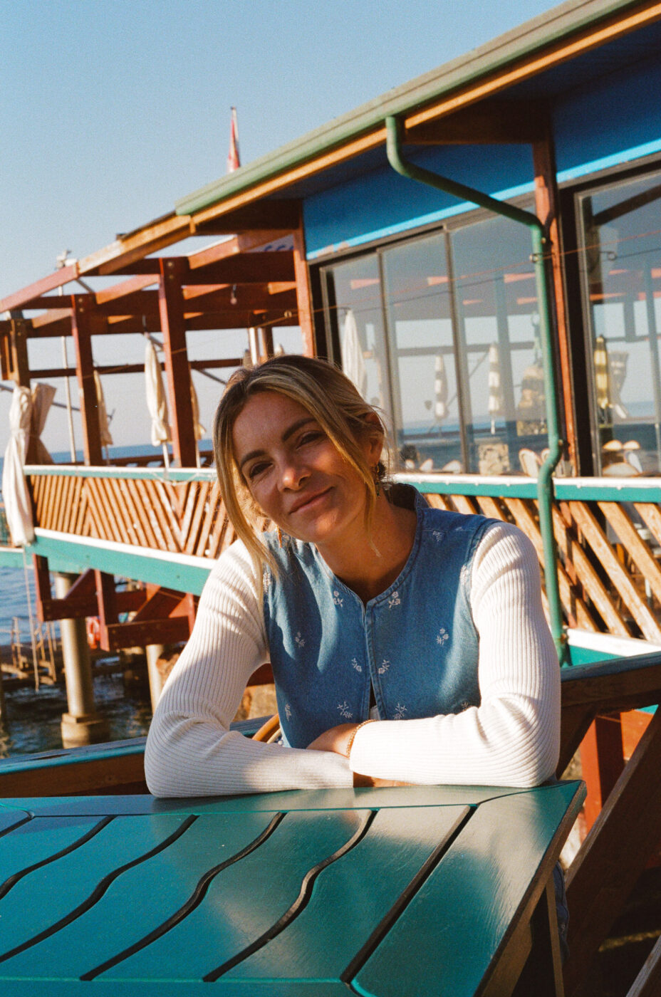 Seaside Smiles - Una donna con i capelli biondi, che indossa un gilet blu su una camicia bianca, siede a un tavolo turchese all'aperto vicino a una struttura in legno sul mare, sorridendo alla fotocamera sotto un cielo limpido e soleggiato.