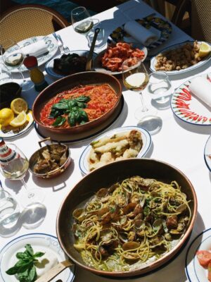 Tavola imbandita per un pranzo all'aperto - Tavola imbandita per un pranzo all'aperto, con piatti di pasta ai frutti di mare, pasta al pomodoro, verdure grigliate, gamberi, carciofi, limoni e bicchieri di vino bianco. La luce del sole illumina la vivace e abbondante tavola.