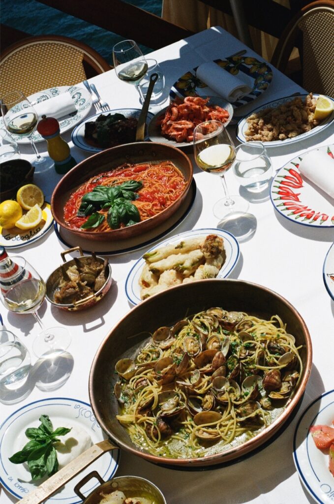 Tavola imbandita per un pranzo all'aperto - Tavola imbandita per un pranzo all'aperto, con piatti di pasta ai frutti di mare, pasta al pomodoro, verdure grigliate, gamberi, carciofi, limoni e bicchieri di vino bianco. La luce del sole illumina la vivace e abbondante tavola.