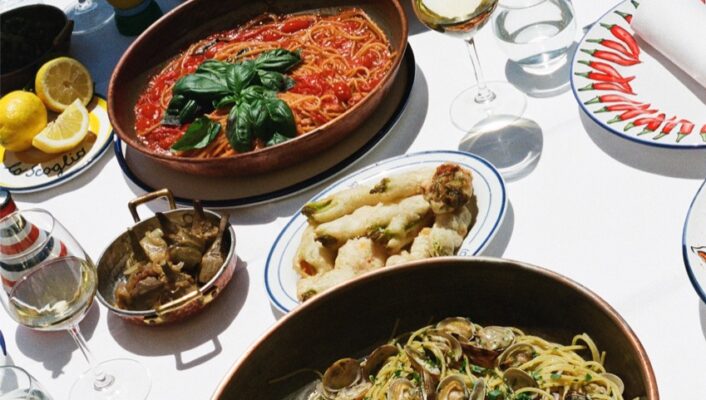 An outdoor table set for a meal, featuring plates of seafood pasta, tomato pasta, shrimp, grilled vegetables, and wine glasses, with sunlight highlighting the colorful dishes and fresh garnishes.