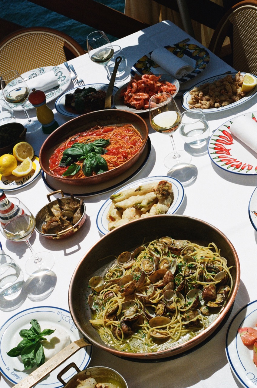 Tavola imbandita per un pranzo all'aperto - Tavola imbandita per un pranzo all'aperto, con piatti di pasta ai frutti di mare, pasta al pomodoro, verdure grigliate, gamberi, carciofi, limoni e bicchieri di vino bianco. La luce del sole illumina la vivace e abbondante tavola.