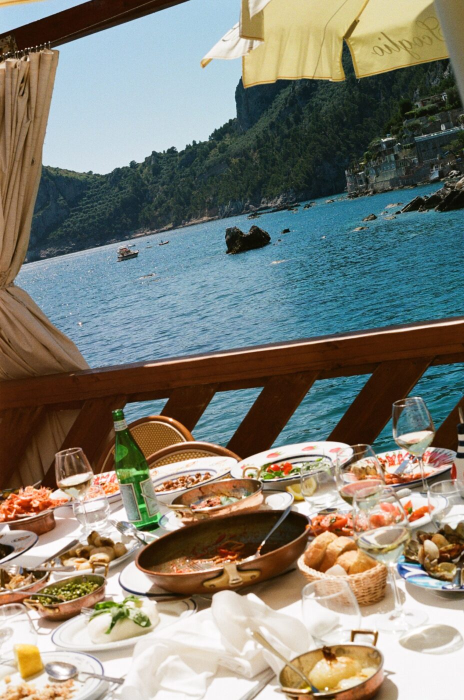 Pranzo di pesce in riva al mare - Una tavola imbandita con piatti di pesce, pane, vino e acqua si affaccia su un mare azzurro e calmo e su una costa rocciosa, con barche in lontananza e ombrelloni gialli in alto. La scena suggerisce un piacevole pasto all'aperto in riva al mare.