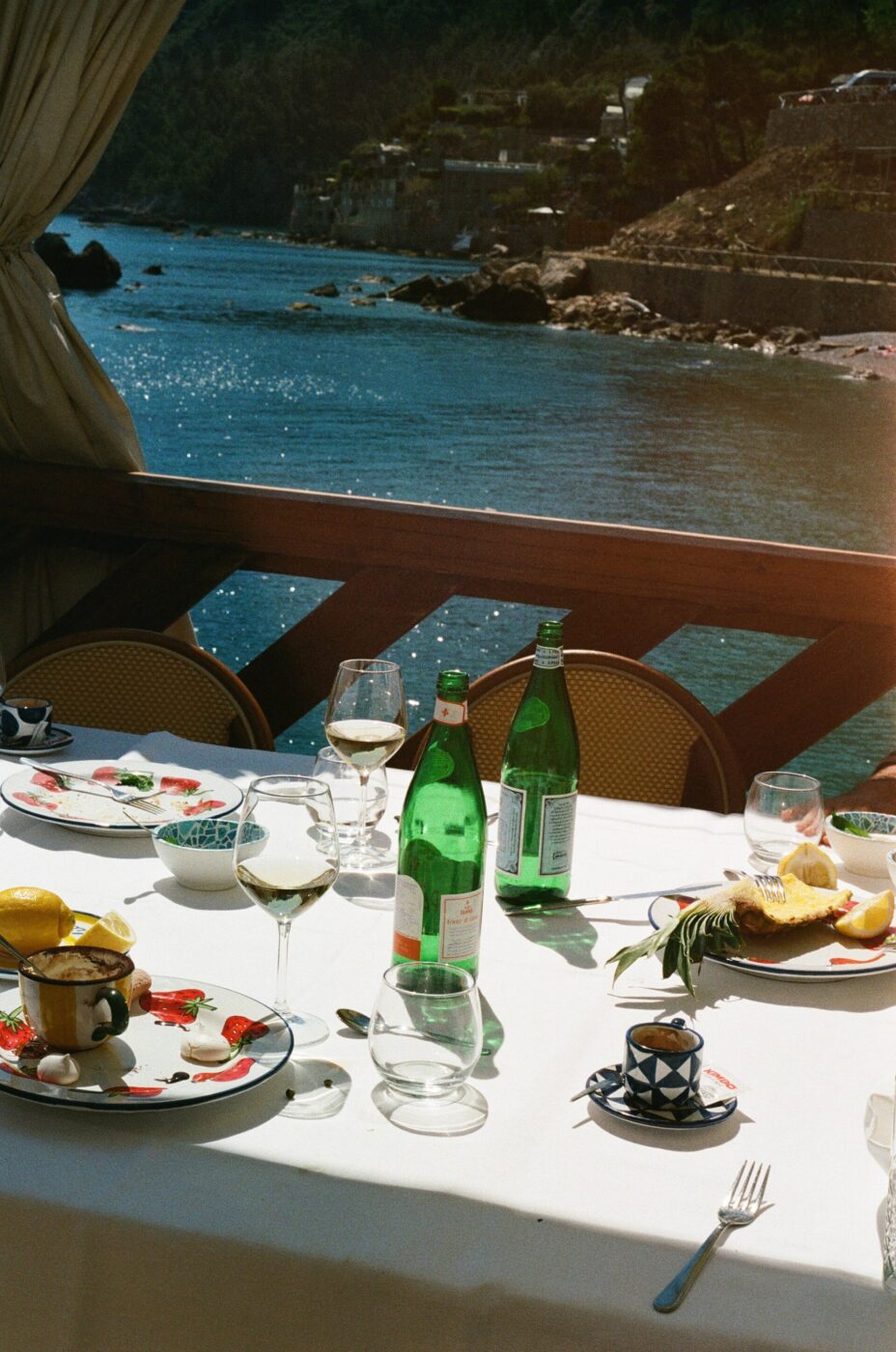 Waterfront Dining - A table set for a meal with bottles of sparkling water, wine glasses, and plates of food overlooks a scenic waterfront with rocky cliffs and blue water in the background.