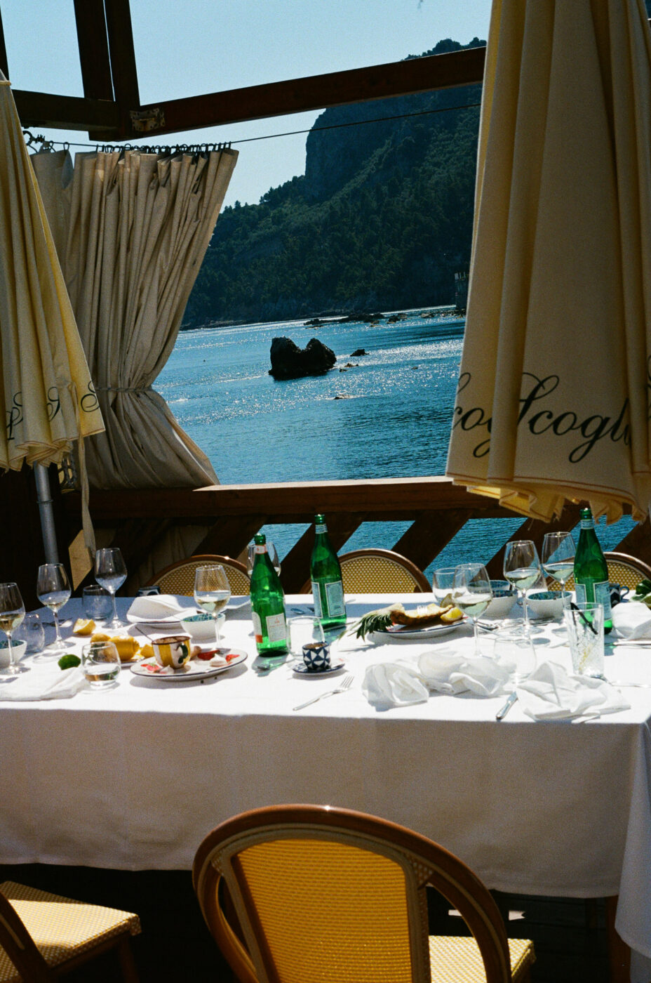 Tavolo del ristorante sul mare - Tavolo del ristorante sul mare con piatti, bicchieri, bottiglie d'acqua verdi e cibo, circondato da sedie di vimini, con vista sull'acqua blu, sulla costa rocciosa e sulla luce del sole che si riflette sul mare.