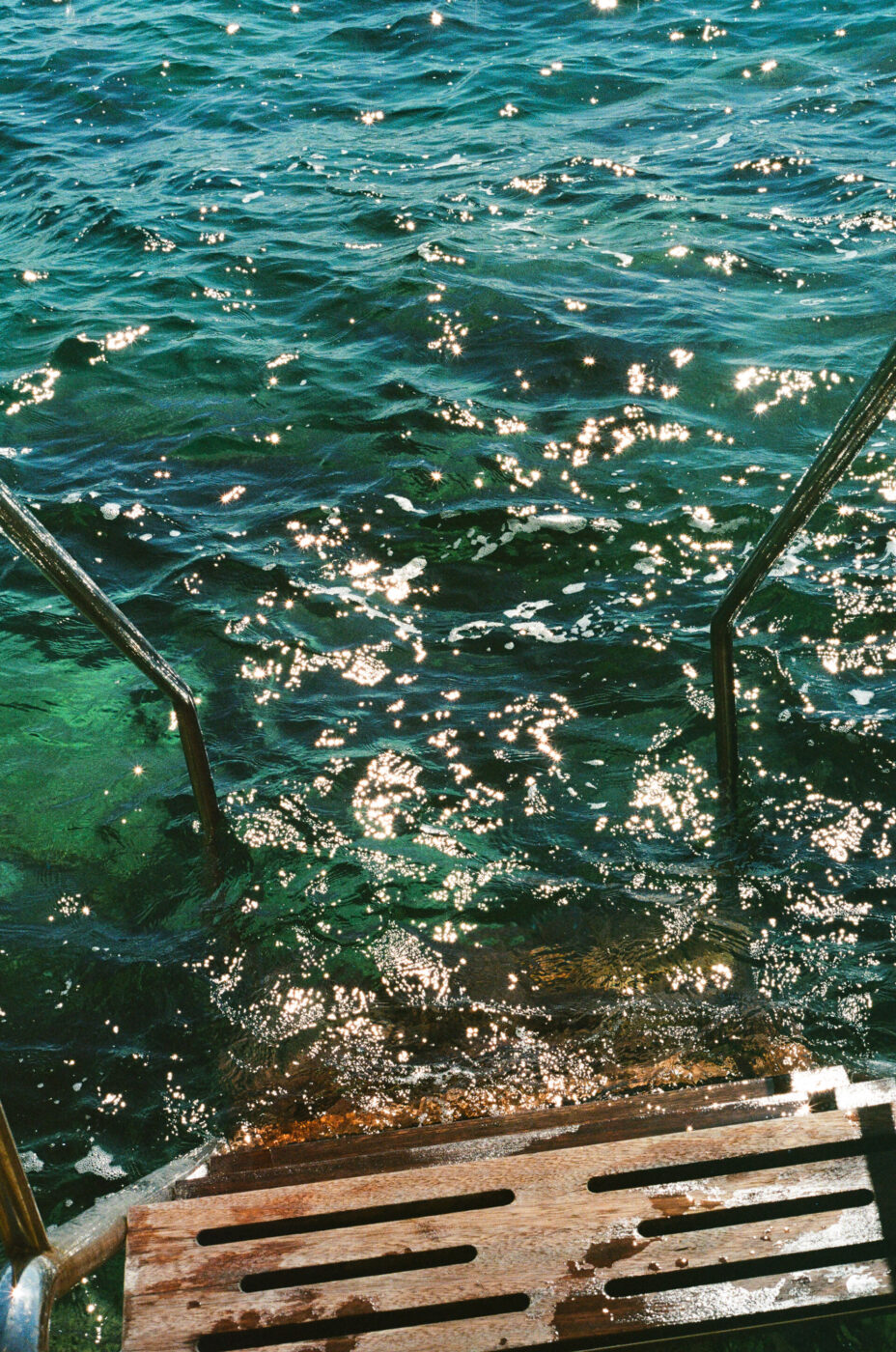 Scala d'acqua turchese - Gradini di metallo scendono in un'acqua turchese, limpida e illuminata dal sole, con riflessi scintillanti sulla superficie. La scena fa pensare a un punto di balneazione o a una scala di attracco che conduce al mare o a un lago.