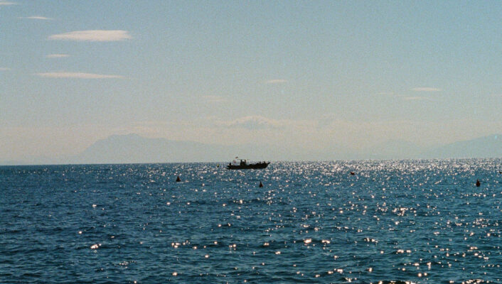 Una piccola barca galleggia su un mare blu scintillante sotto un cielo limpido, con le montagne lontane leggermente visibili all'orizzonte.